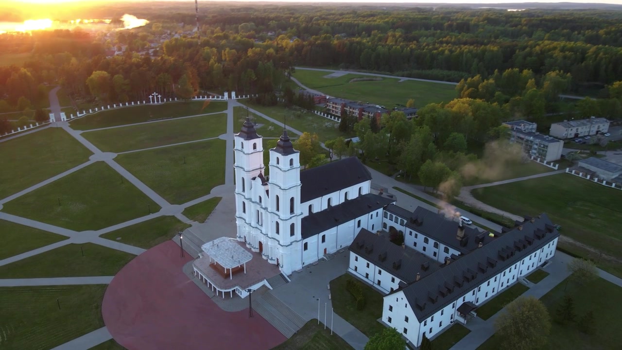 Drohnenaufnahme über der Basilika Mariä Himmelfahrt in Aglona, Lettland. Fängt dramatische Lens Flare vom Sonnenuntergang und aufsteigenden Schornsteinrauch vom historischen Gebäude ein.