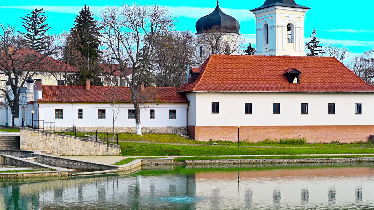 Objevte krásnou historickou stavbu nacházející se u klidného jezera v Rumunsku. Scenérie ukazuje přírodu ožívající na začátku jara s jasnou oblohou a svěží zelení.