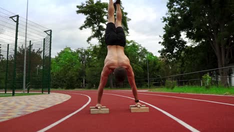 the young athlete is standing upside down leaning on special stands on race track outdoors. young man topless in black shorts