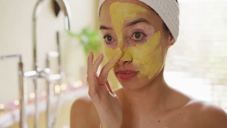 portrait of biracial woman looking into mirror and applying face mask