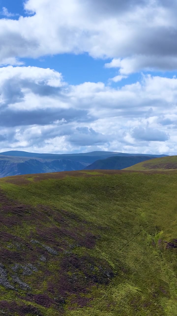 La telecamera panoramica su colline erbose e coperte di erica delle Highlands scozzesi sotto la luce del giorno intensa e nuvole sparse.