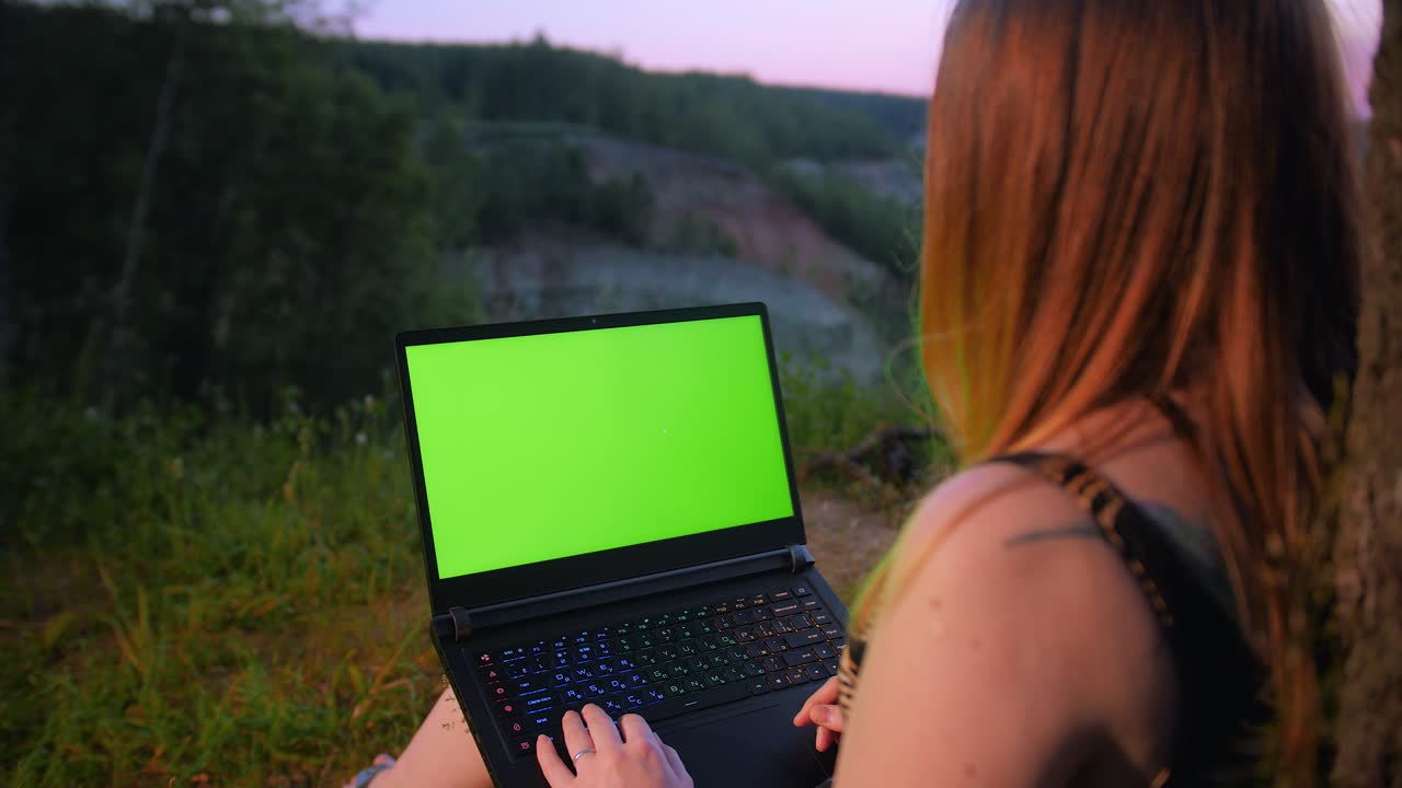 una joven trabaja en una computadora portátil con una pantalla verde cerca de un árbol en la cima de una montaña.