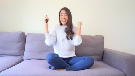 Mujer-Asiática-Feliz-Viendo-Noticias-En-La-Televisión-Con-Mando-A-Distancia-Y-Celebrando-Con-Gesto-De-Manos,-Lotería-O-Deporte-Ganador,-Cámara-Lenta-De-Cuadro-Completo