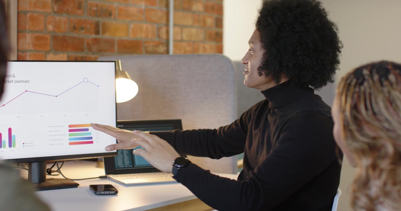 African American woman turning to monitor at desk after coworker pointing, explaining chart trends