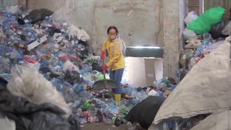 A-Girl-With-Dreadlocks,-In-Yellow-Boots-In-Protective-Mask,-Scoops-Used-Bottles-With-A-Large-Duck-Shovel-At-A-Plastic-Recycling-Factory