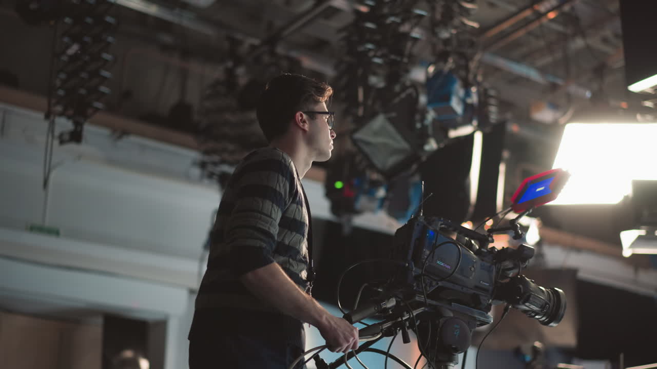Camera operator adjusting broadcast camera rig on dolly in modern television studio with overhead lighting rigs and cables, focusing on shot composition and adjustment during live production