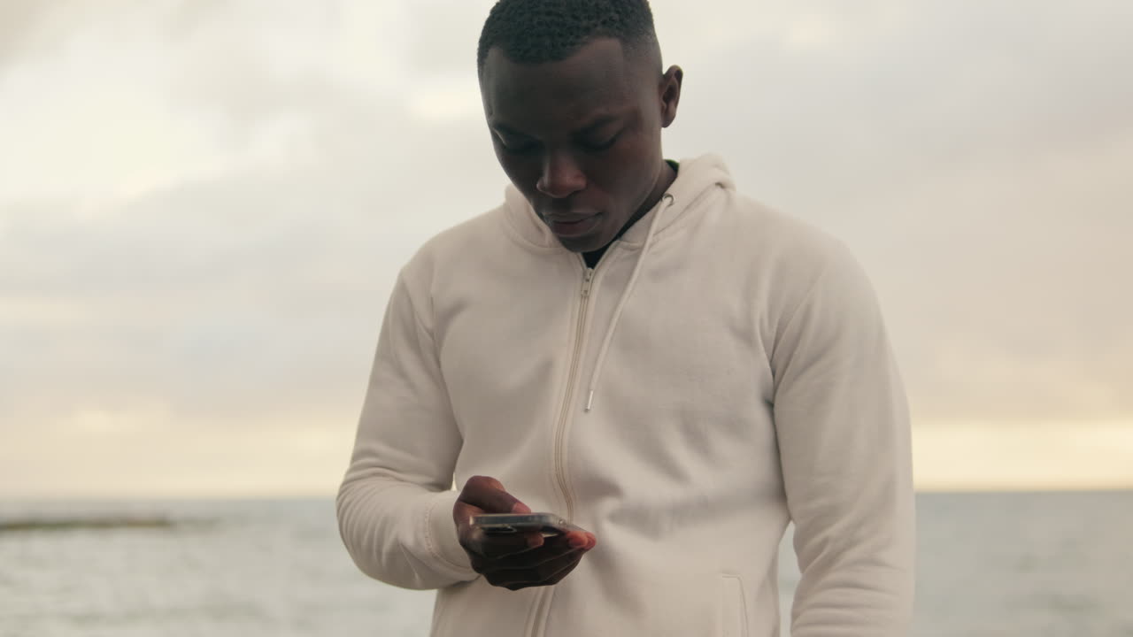Ung sort mand bruger smartphone udendørs ved havet