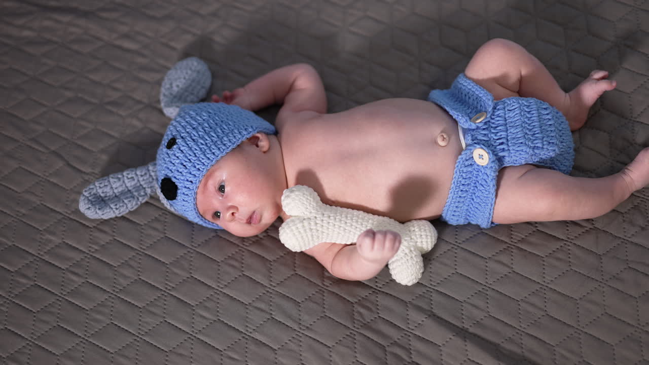 Infant in a blue knitted costume lying on the bed. White knitted toy bone near baby's hand. Sweet baby on the grey background.