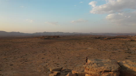 Vista-Aérea-Del-Hombre-Parado-En-La-Cima-De-Una-Colina-Rocosa-Y-Mirando-El-Sol-Del-Atardecer-Sobre-El-Paisaje-Seco-Del-Desierto-De-Utah-Usa