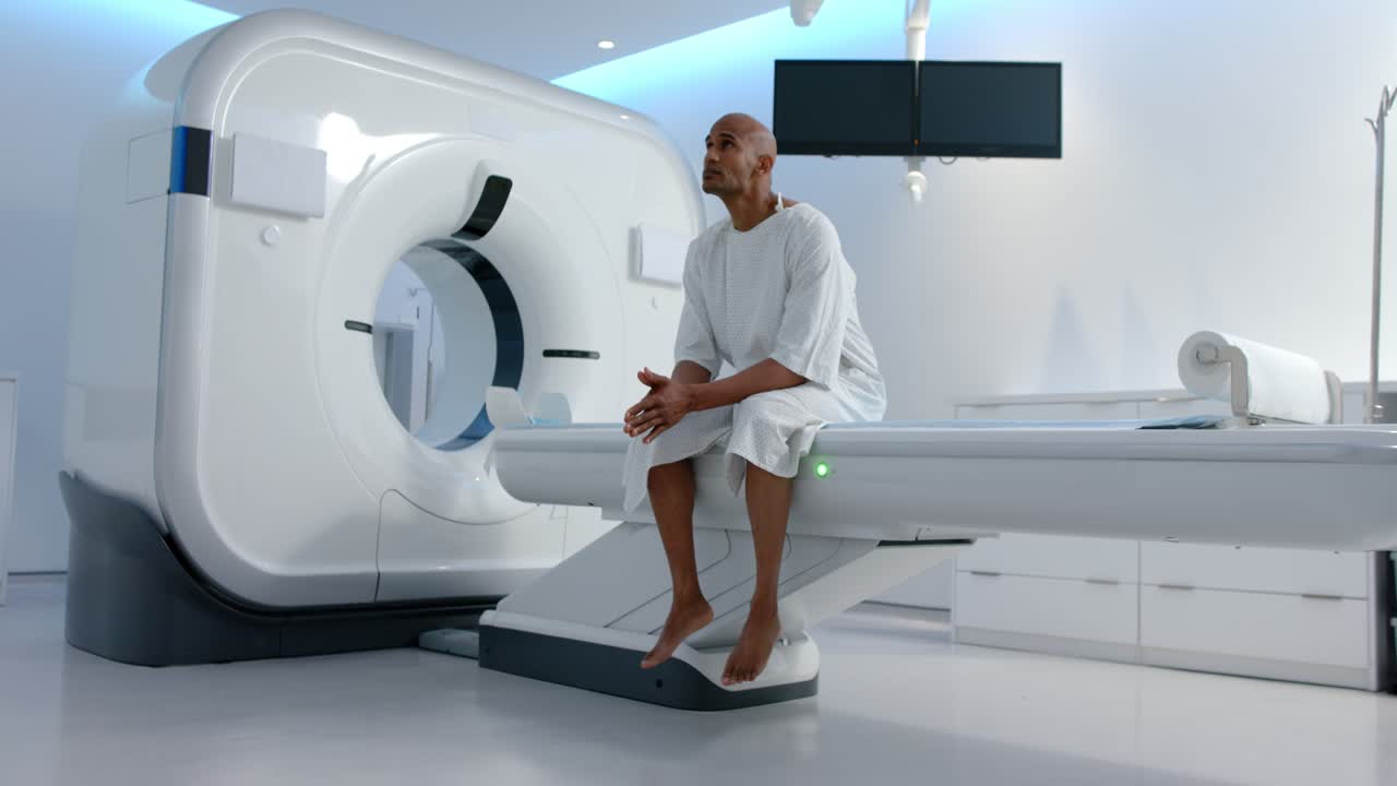 Uomo afroamericano in camice ospedaliero seduto su un tavolo nella sala scanner, che osserva una luce verde sul portale.