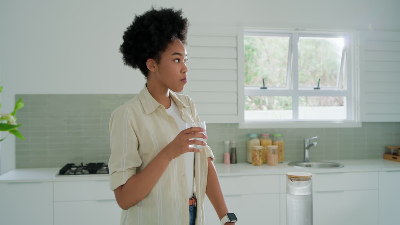 Donna afroamericana sui vent'anni che ha sete, versa da una caraffa e sorseggia da un bicchiere all'isola della cucina