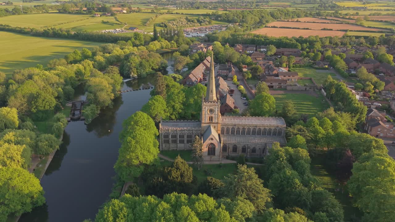 Toma de dron de ángulo alto mirando hacia la gótica Iglesia de la Santísima Trinidad, inclinándose hacia arriba para revelar el extenso paisaje primaveral y el horizonte de Stratford-upon-Avon.