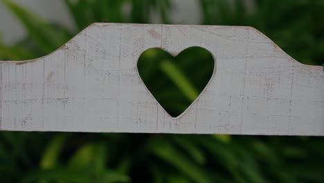 White-wooden-bench-with-a-heart-shaped-cutout-against-a-green-leafy-background