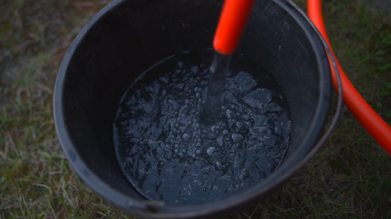 Nærbillede af en sort spand, der står på græs, mens en vandstråle hældes i fra en orange slange, der skaber krusninger og plask i naturligt dagslys