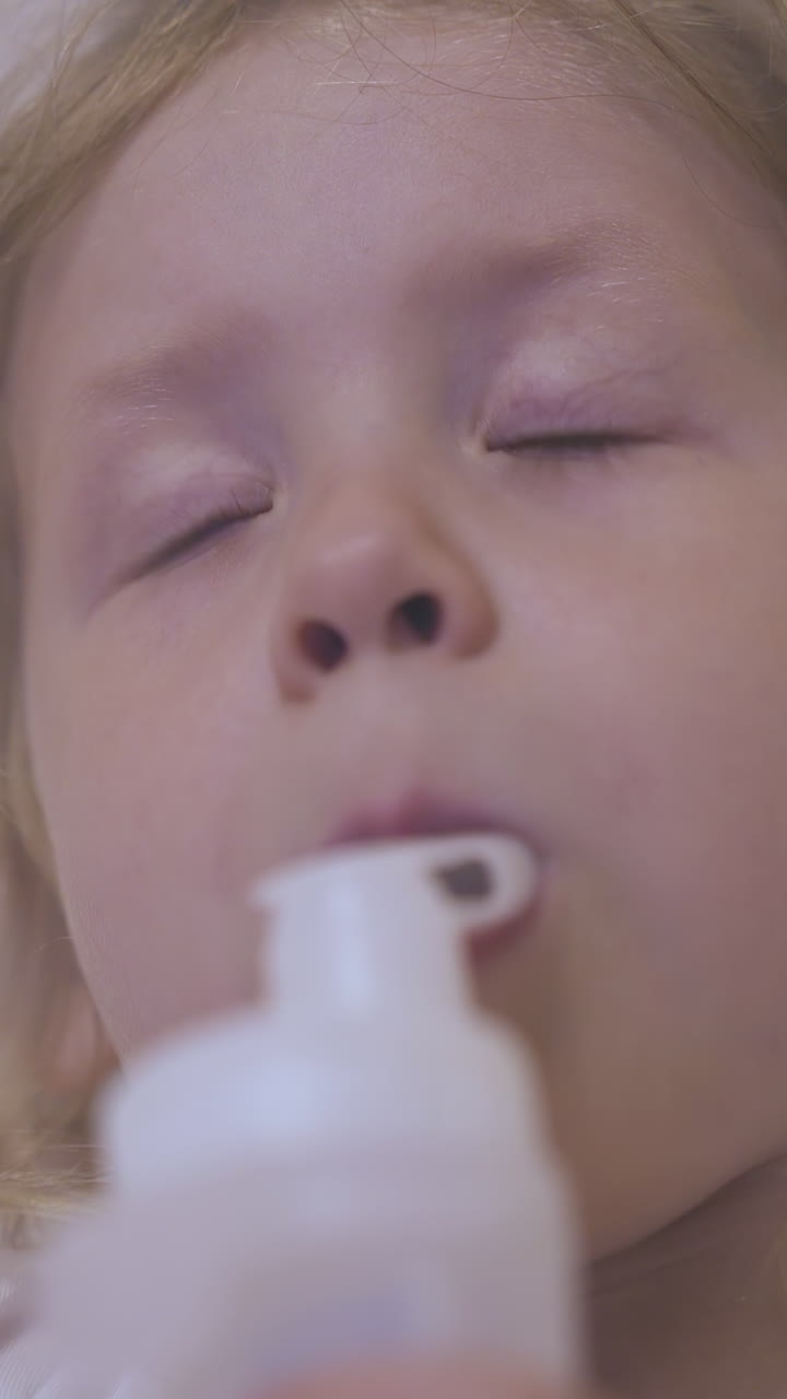 sad small girl breathes with modern inhaler lying in comfortable bed near beige wall in light room extreme close view