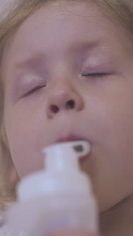sad small girl breathes with modern inhaler lying in comfortable bed near beige wall in light room extreme close view