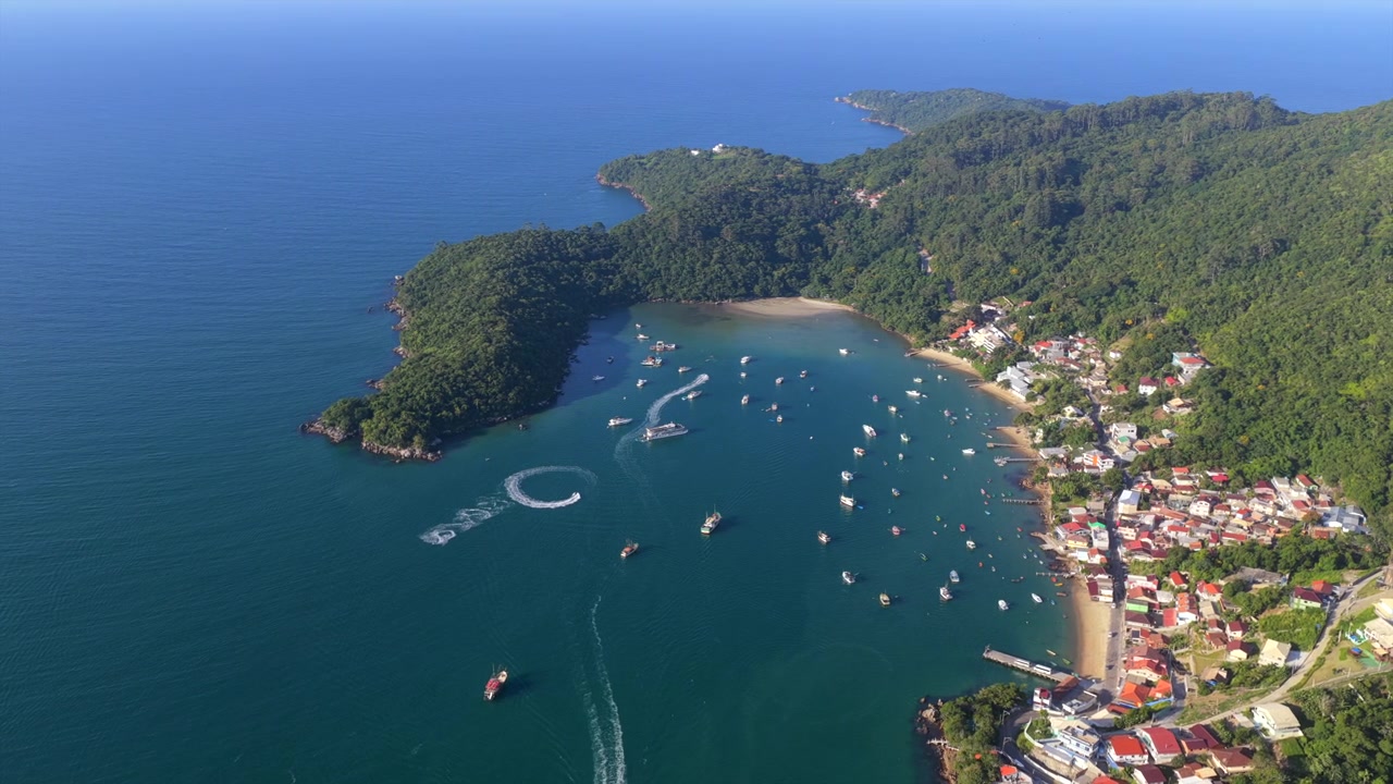 Barcos ancorados nas águas turquesa da Praia de Caixa D'aço, cercados por vegetação exuberante e uma cidade costeira. Ideal para promoções de viagens ou documentários.