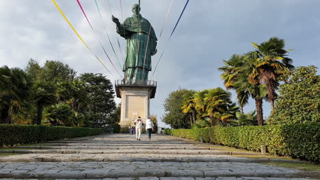 Couple-walk-on-stairs-towards-giant-statue-of-San-Carlo-in-Arona,-Italy