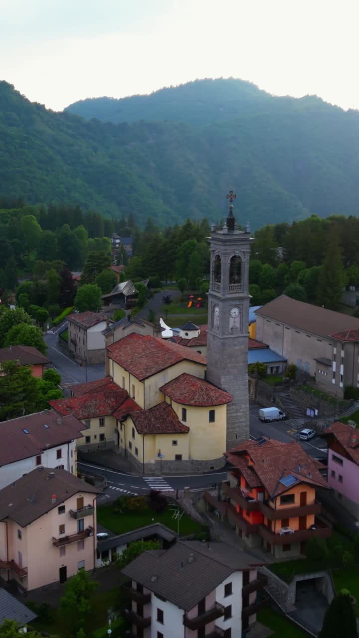 Vista drone della chiesa del villaggio di Selvino, con un campanile prominente e architettura tradizionale. Circondata da edifici residenziali e montagne verdi e rigogliose, questa scena cattura il sereno fascino