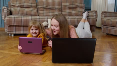 mother and small daughter child sit on warm wooden floor at home working on laptop, watching cartoon