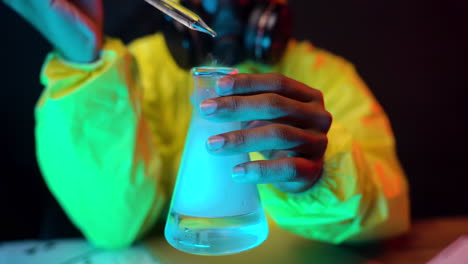 científico de traje amarillo hazmat sostiene un recipiente de vidrio de laboratorio que deja caer líquido transparente mientras usa una máscara de gas respiratorio, experimento peligroso cerca de las manos de trabajo del químico