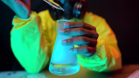 yellow hazmat suit scientist hold laboratory glass container dropping transparent liquid while wearing respiratory gas mask, dangerous experiment close up of chemist working hands