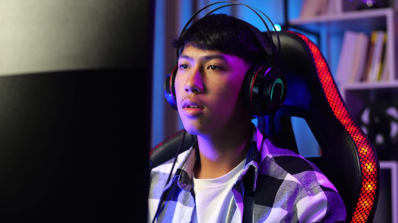 Primo piano di un giovane streamer asiatico che gioca online al computer personale, in un videogioco in live streaming dove sfida e minaccia i concorrenti, sconfiggendoli. La scrivania è illuminata da una striscia LED RGB.