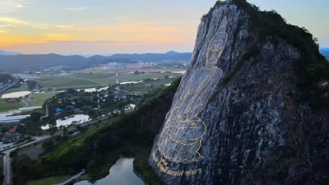 4K-Drohnenaufnahmen,-Die-Ein-Bild-Von-Buddha-Umkreisen,-Der-Im-Schneidersitz-Sitzt-Und-Mit-Gold-In-Die-Nordwand-Eines-Kalksteinhügels-In-Khao-Chi-Chan,-Thailand,-Eingraviert-Ist