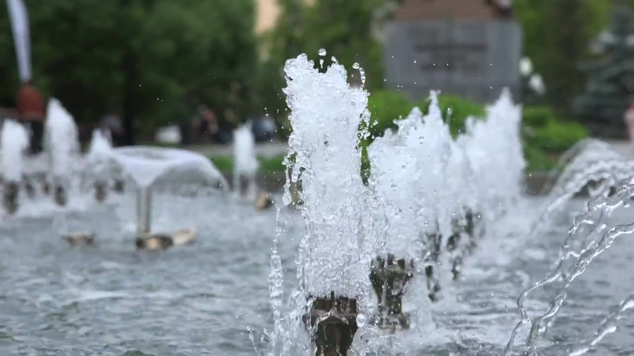Un haut jet d'eau central s'élève au-dessus de buses basses, projetant un épais panache blanc sur l'eau ondulante du bassin, sous une douce lumière du jour, avec des arbres verts flous, des tons froids et une atmosphère de parc public calme