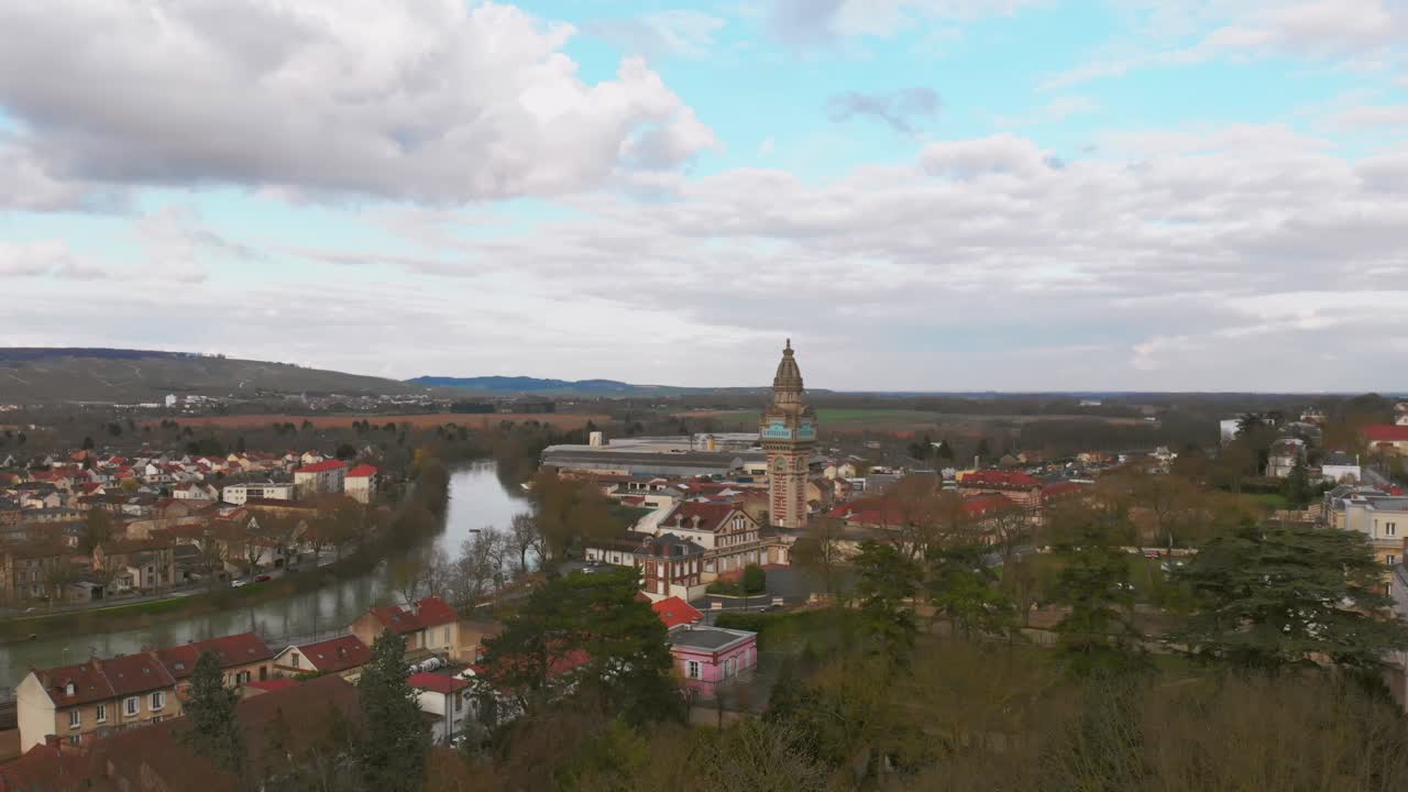 Aerial dolly down revealing the town of Champagne, France, with a river, church tower, and rolling countryside under cloudy skies