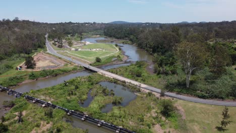 Drone-Volando-Hacia-Un-Parque-Verde-Sobre-Una-Carretera,-Un-Oleoducto-Y-Un-Puente-Sobre-Un-Río-Recientemente-Inundado