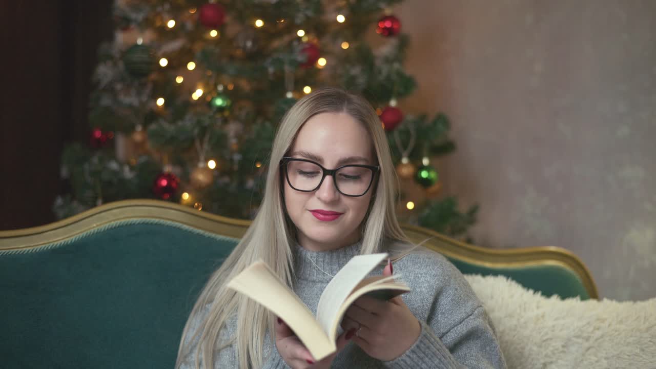 A young woman smells the scent of a book while sitting on a green sofa near a beautifully decorated Christmas tree. Warm lights and festive ornaments create a cozy and peaceful holiday ambiance