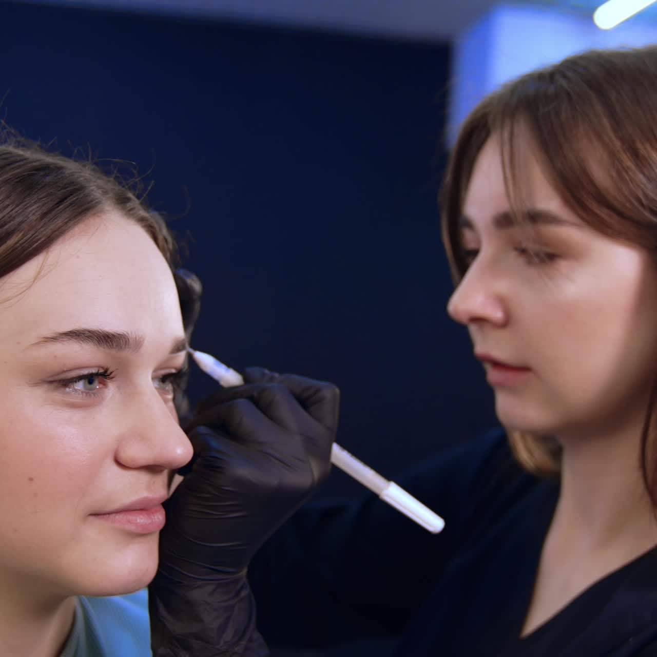 Young beautiful Caucasian girl is at salon. Brunette long-haired brow master drawing lines for future eyebrows. Close up