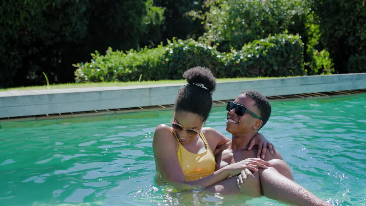 Pareja afroamericana, hombre levantando a la mujer, acunándola y mostrando afecto en la piscina del jardín con gafas de sol.