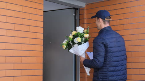 woman opening entrance door, receiving flowers from courier and signing paper