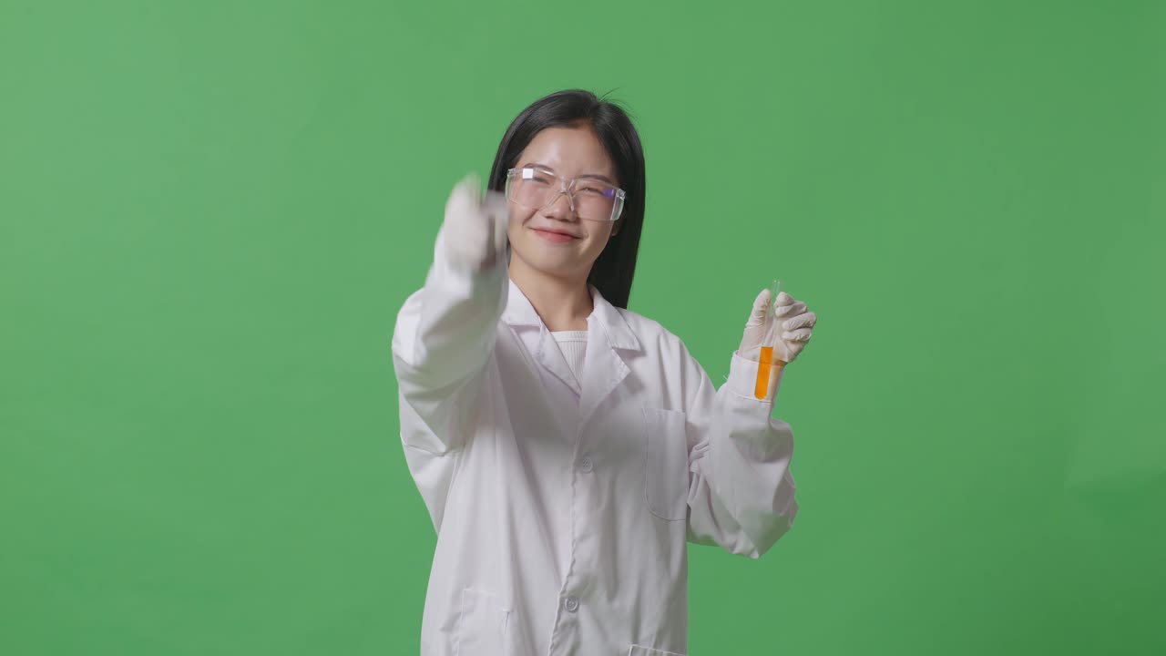 실험 튜브에 오렌지색 액체를 넣은 아시아 여성 과학자가 웃으며 가을 만지고 실험실의 녹색 스크린 배경에 서있는 동안 당신을 가리키고 있습니다.