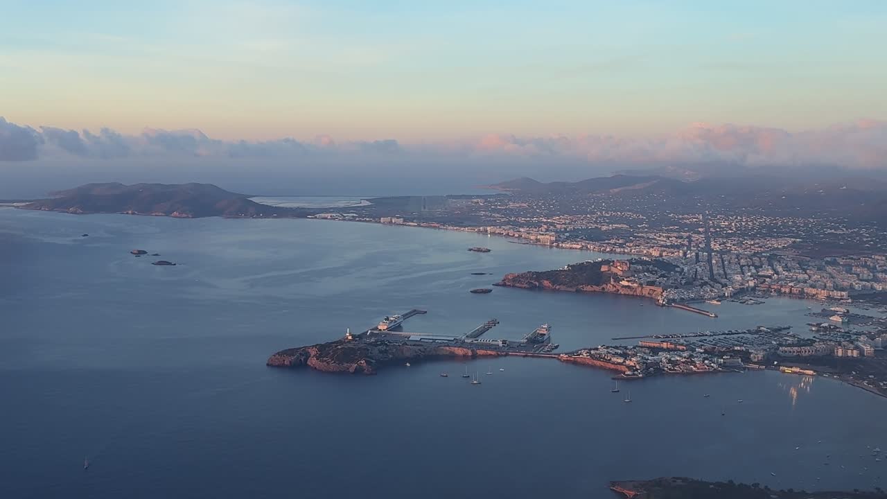 An eleveted right side aerial view of Ibiza city center, harbor, and the airport runway ahead, illuminated by the sunrise sun, taken from an airplane cockpit. Handheld camera cockpit shot
