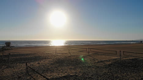 Wide-flyover-empty-volleyball-courts-and-nets-past-lifeguard-tower-to-beautiful-ocean-sunset-and-the-surf-with-aerial-4k-drone-in-Huntington-Beach,-Southern-California