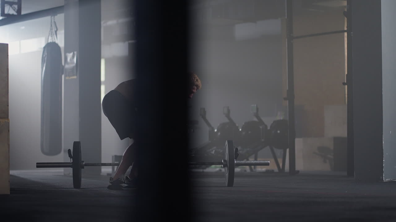슬로우 모션으로 셔츠를 입지 않은 젊은 남자가 배경 조명에서 체육관에서 바벨을 넘어 버피를 점프합니다.