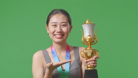 close up of asian woman with a gold medal pointing to a gold trophy in her hand and smiling being proud winning as the first winner on green screen background in the studio