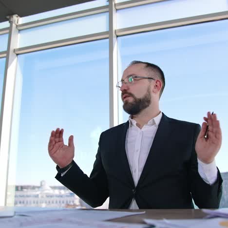 Successful businessman leading a conference. Male entrepreneur in glasses and costume sitting at the table and talking to someone in light office