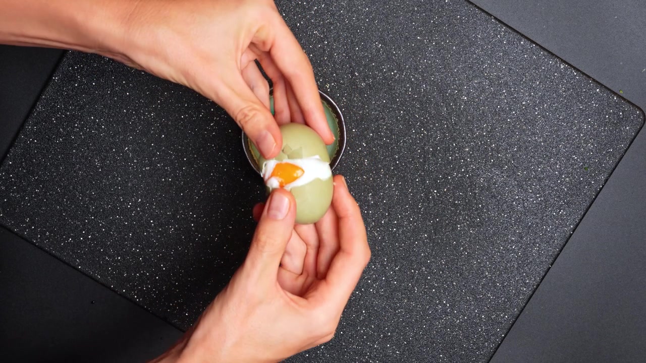 Hands prepare a green egg on a black cutting board in a kitchen setting. Hands hold a green egg above a black cutting board in a kitchen.