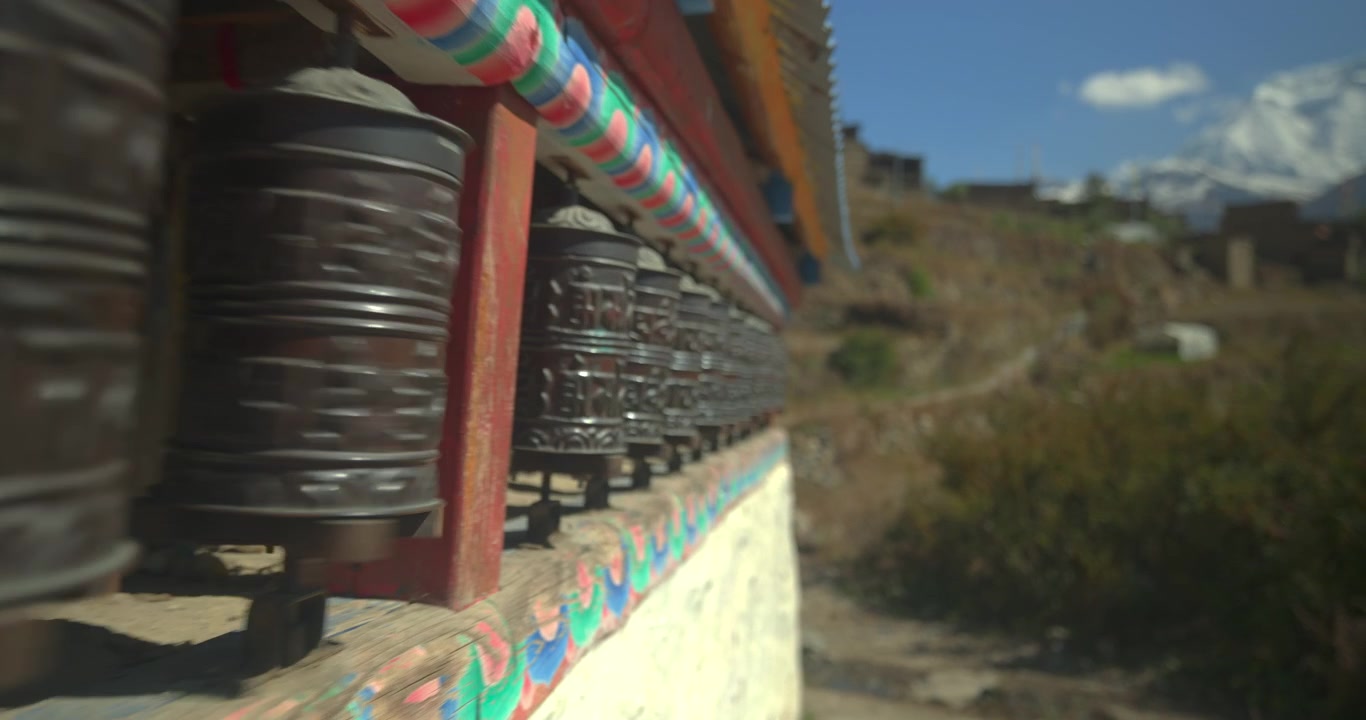 Circuito dell'Annapurna, Manang, Nepal, ripresa scorrevole lungo una fila di tradizionali ruote di preghiera buddiste in metallo installate su un muro di pietra dipinto nel villaggio di Manang