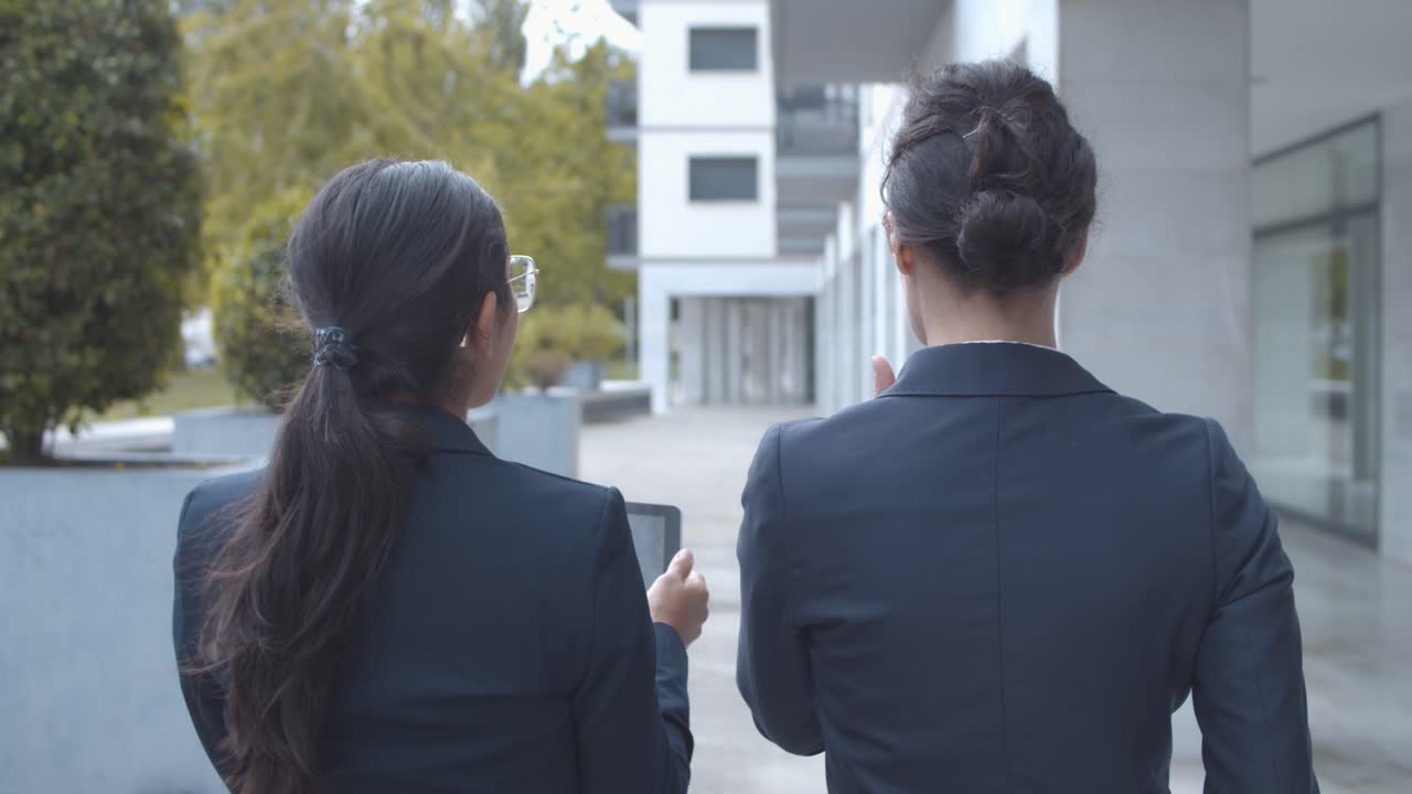 두 명의 여성 관리자가 사무실 건물에서 걷는 동안 프로젝트에 대해 토론하고 태블릿을 사용하는 모습