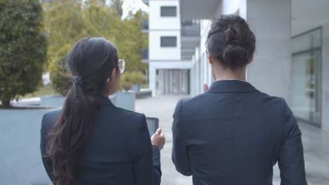 back view of two female managers discussing project and using tablet while walking at office building