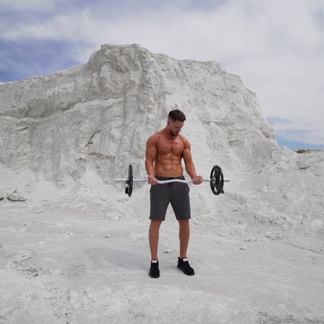 young guy in shorts trains his muscular body outdoors. bodybuilder lifting metal barbell in white mountains. slow motion.