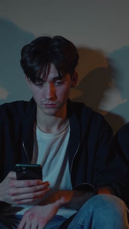 young man sitting on comfortable sofa, checking smartphone with serious expression in dimly lit room, revealing digital connection and solitary moment of modern lifestyle