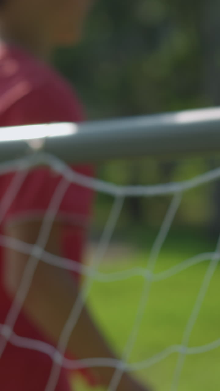A blurred view of a goalkeeper in red attempts to save a ball shot into the net in a grassy field