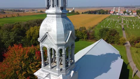 Drone-shot-strafing-over-a-community-chapel,-showing-the-church-bells,-churchyard,-and-the-surrounding-farms,-located-in-Montreal,-Quebec-in-Canada