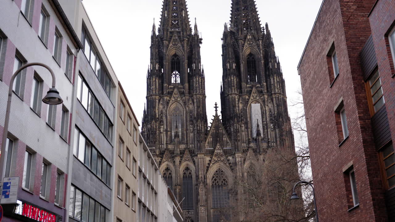 A striking perspective of the majestic Cologne Cathedral spires framed by contemporary urban buildings. The juxtaposition highlights the timeless beauty of Gothic architecture amidst modern city life.